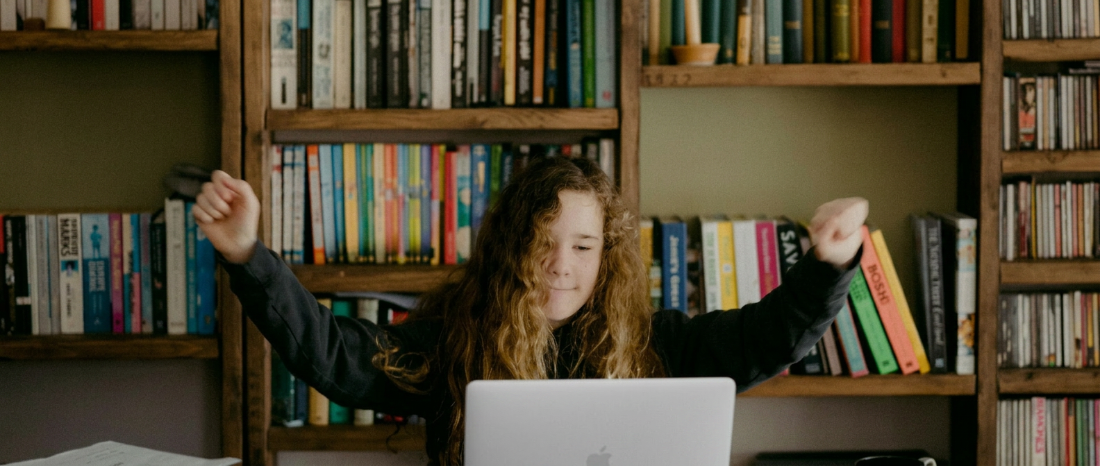 A teenage student celebrating a breakthrough at her laptop in front of a wall of books